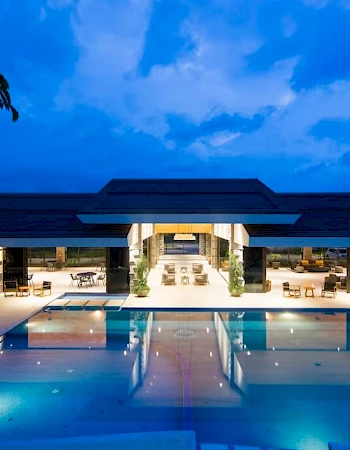 A luxurious modern building with an illuminated outdoor swimming pool at dusk, surrounded by lounge seating and a scenic evening sky.