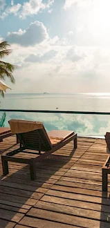 Poolside view with lounge chairs, umbrellas, and palm trees overlooking a tranquil ocean under a partly cloudy sky.