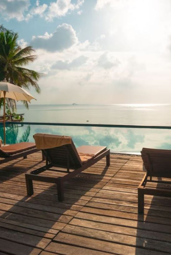 Lounge chairs and umbrellas sit by a serene pool overlooking the ocean, with palm trees and a sunlit sky in the background.