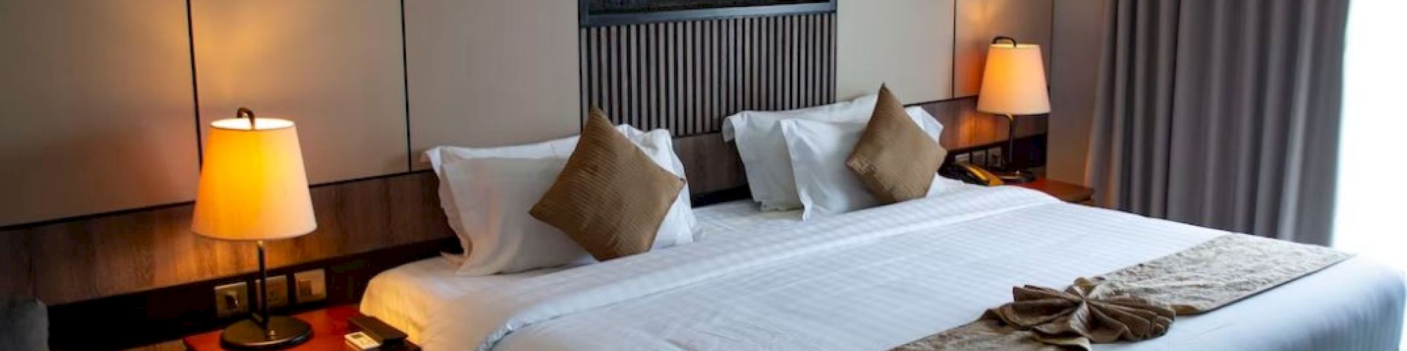 A modern hotel room features a large bed with pillows, nightstands, lamps, and a wall-mounted decor above the headboard.