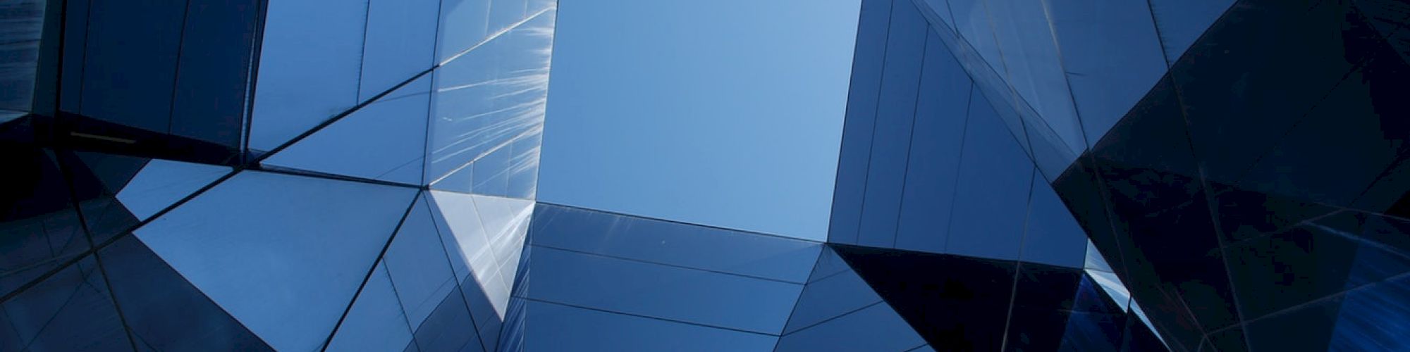 The image shows a geometric, abstract view of a modern glass building, with reflective surfaces converging to a clear blue sky above.