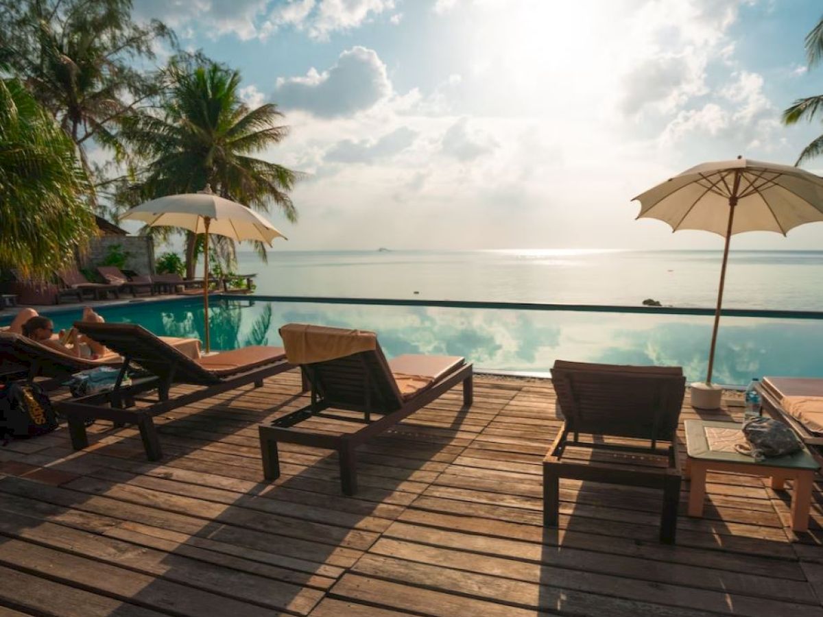 A tranquil poolside scene with lounge chairs, umbrellas, and palm trees overlooking a serene ocean view under a partly cloudy sky.