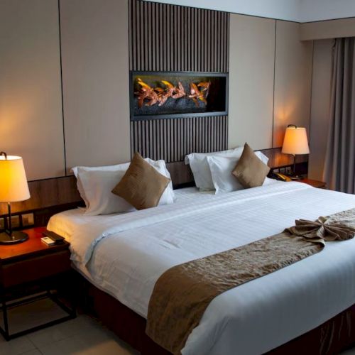 A modern hotel room with a large bed, pillows, lamps on side tables, soft lighting, and contemporary decor, including a wall-mounted art piece.