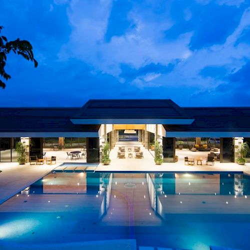 A luxurious modern building with expansive glass windows is illuminated at night, overlooking a large, well-lit swimming pool.