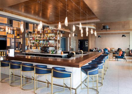 A stylish bar with a wooden counter, modern lighting, and blue stools, surrounded by shelves of bottles, offers a chic lounge atmosphere.