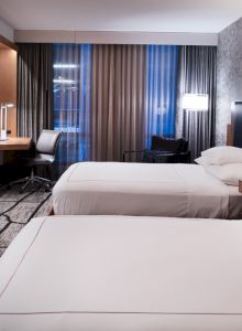 The image shows a modern hotel room with two beds, a desk, a TV, and stylish decor, including patterned wallpaper and curtains.