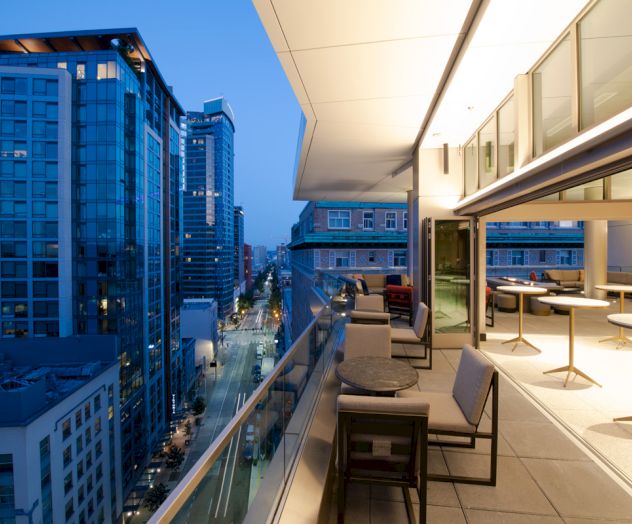 A modern urban balcony at dusk with seating, overlooking a city street and high-rise buildings, lit gently by building lights.