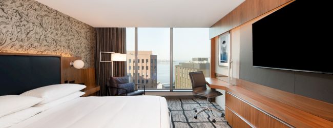 A modern hotel room with a large bed, wall art, desk, TV, and city view through a window.