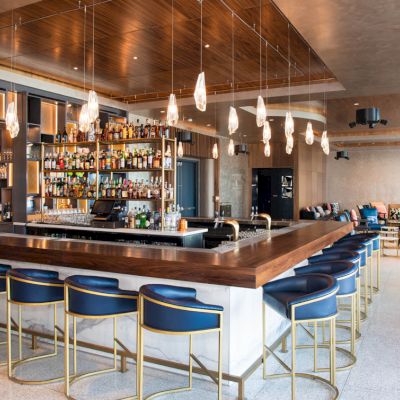 A modern bar with a central counter, stylish seating, and hanging pendant lights in a spacious, contemporary lounge setting.
