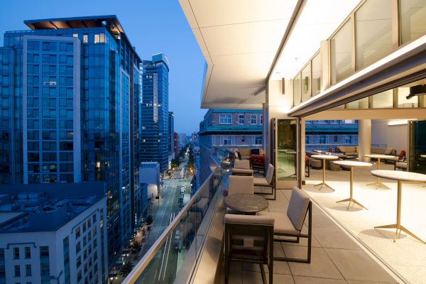 A modern cityscape at dusk with tall buildings, a stylish outdoor terrace with seating, and city lights illuminating the street below.
