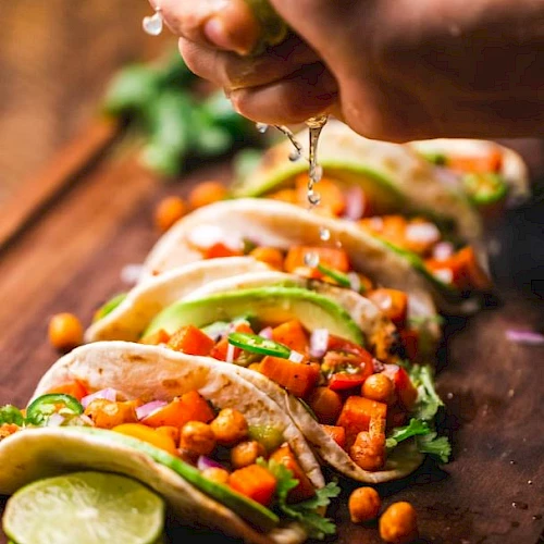 Tacos filled with chickpeas, avocado, and veggies, placed on a board with lime being squeezed over them.