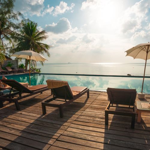 Sunlit poolside deck with lounge chairs and umbrellas overlooking a serene water view, flanked by palm trees under a cloudy sky.