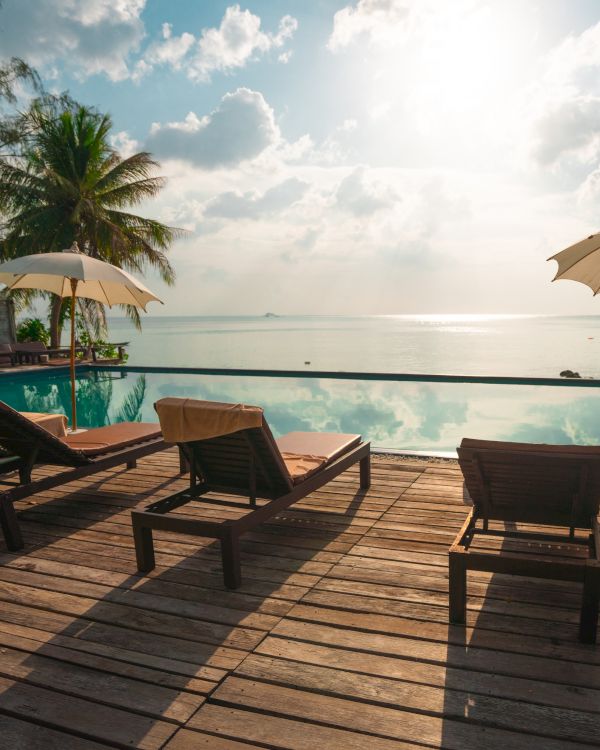 Sunlit poolside deck with lounge chairs and umbrellas overlooking a serene water view, flanked by palm trees under a cloudy sky.