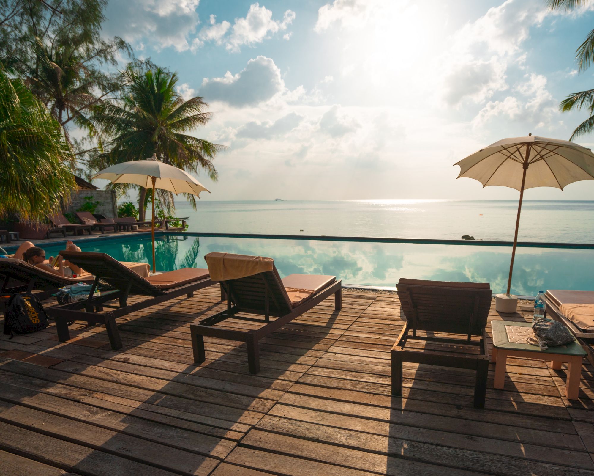 Wooden deck with lounge chairs and umbrellas facing an infinity pool, overlooking the ocean under a bright sky.