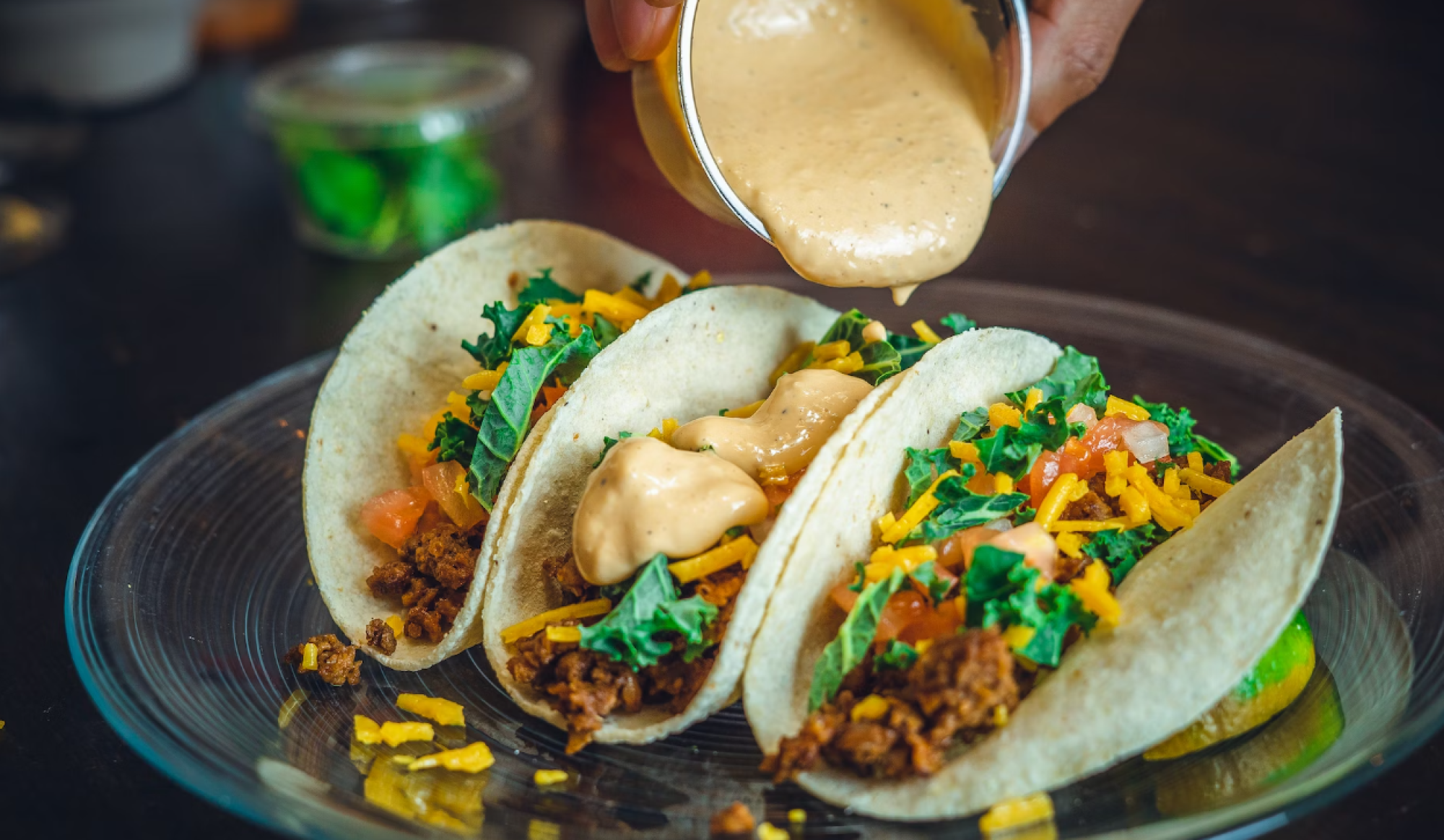 Three tacos with toppings like meat, cheese, lettuce, tomato, and sauce being poured over them on a clear plate.