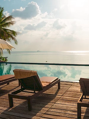 Lounge chairs and umbrellas line a wooden deck by an infinity pool, overlooking a serene ocean view with palm trees and a bright sky.