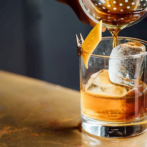 A cocktail being poured into a glass with ice and a citrus garnish clipped to the rim, on a bar counter.