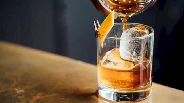 A cocktail being poured into a glass with ice and a citrus garnish clipped to the rim, on a bar counter.