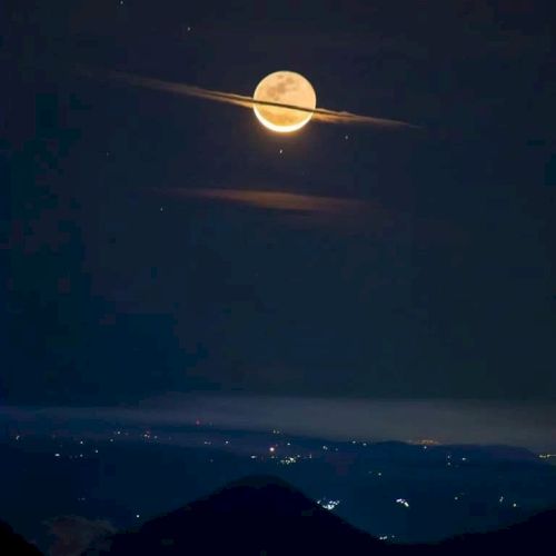 A full moon with cloud rings, resembling Saturn, shines over a dark, mountainous landscape dotted with distant city lights.
