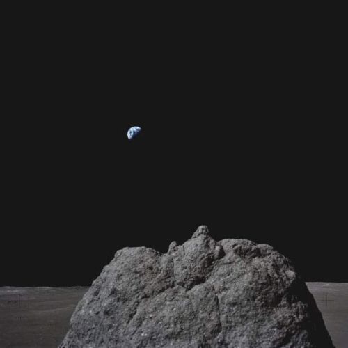 A lunar surface with a large rock is in the foreground, and Earth appears in the distant dark sky.