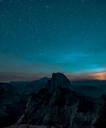 A dark, mountainous landscape under a starry night sky, with a glowing horizon peeking through clouds.