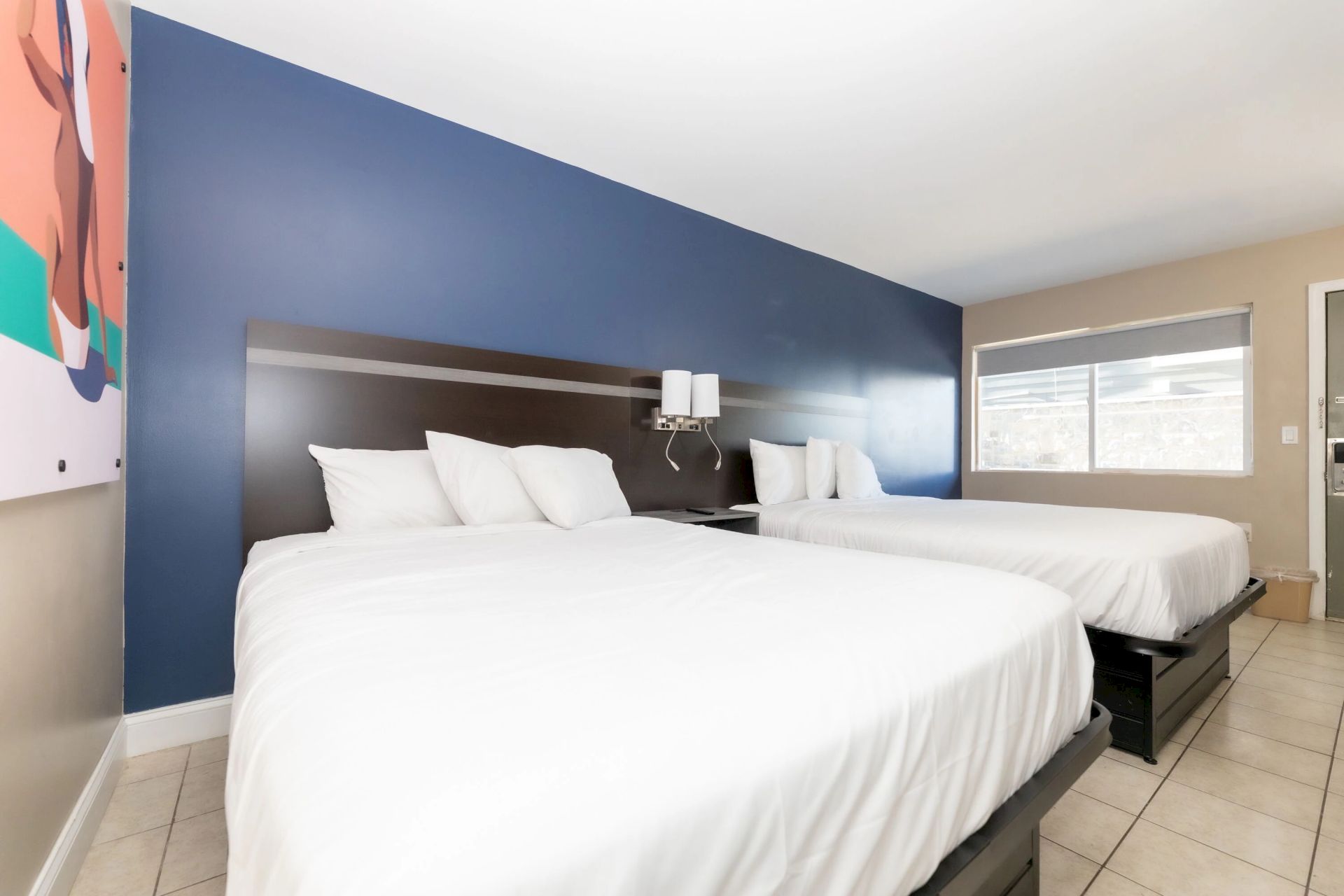 The image shows a hotel room with two neatly made beds, a blue accent wall, a painting, and a window with blinds.