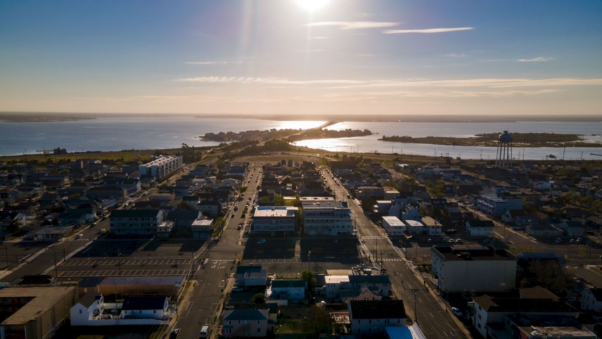 Aerial view of a coastal town at sunset, with streets, buildings, and water in the background under a bright sky.