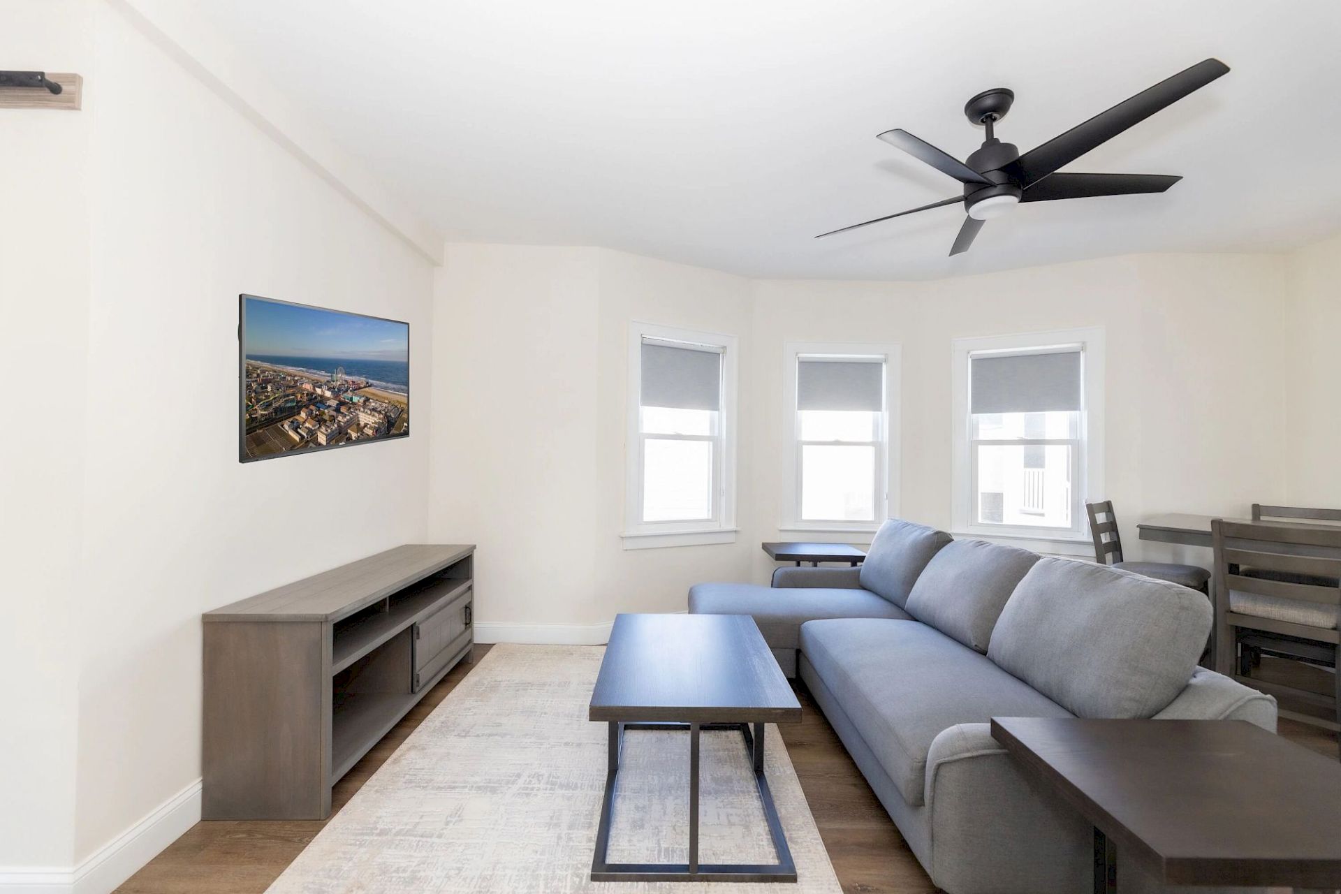 A modern living room with a gray sectional sofa, wall-mounted TV, coffee table, ceiling fan, and three windows with blinds.