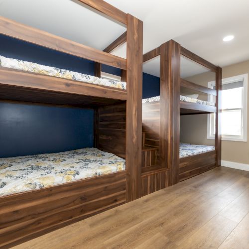 A room with wooden bunk beds, floral bedding, a pillow, a window, and an air conditioner, all on a wooden floor.