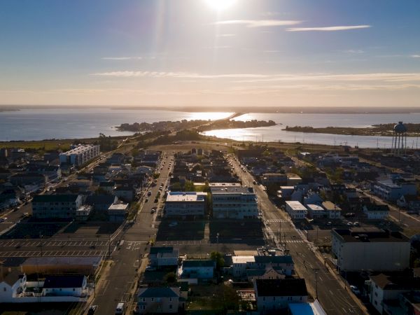 Aerial view of a coastal town at sunset, with buildings, streets, and water in the background under a bright sun.