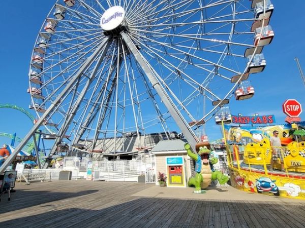 A large Ferris wheel dominates the amusement park scene with colorful stalls and rides beneath, under a clear blue sky.
