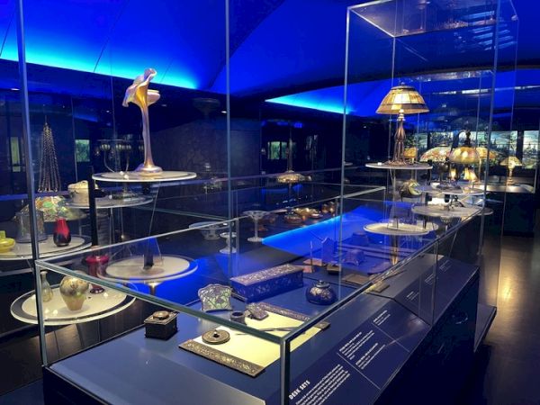 The image shows a museum display with glass cases holding various art objects and lamps, under blue ambient lighting.