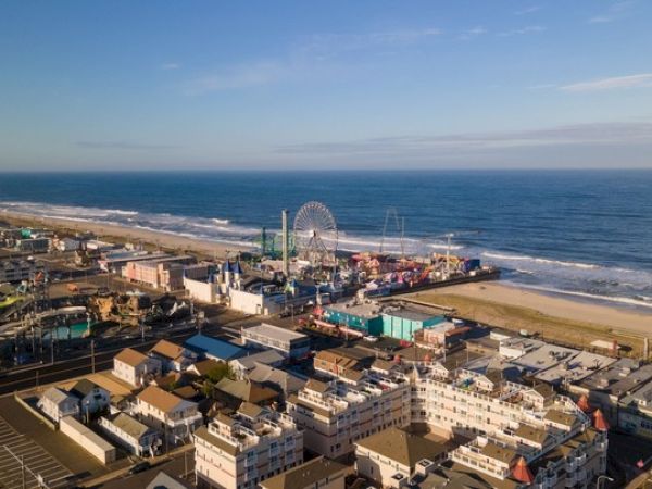 Aerial view of a beachfront amusement park with a Ferris wheel, surrounded by buildings and the ocean under a clear sky.