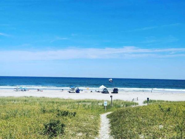 A sandy path leads through grassy dunes to a beach with people, umbrellas, and the ocean under a clear blue sky.