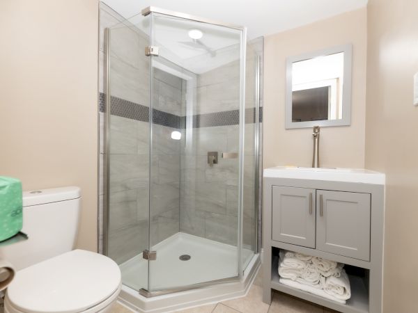 The image shows a modern bathroom with a glass shower, a toilet to the left, and a sink with a mirror and towels beneath.