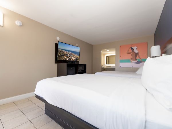 The image shows a hotel room with a TV, white bed, wall art, and a bathroom in the background.