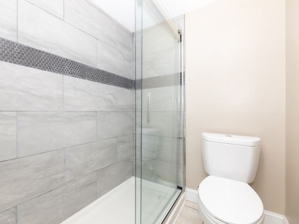 A bathroom with a walk-in glass shower and a toilet, featuring light gray tiles with a decorative dark accent stripe.