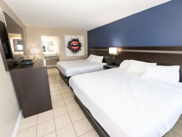 The image shows a hotel room with two beds, a wall-mounted TV, and a striking painting of red lips on the wall.
