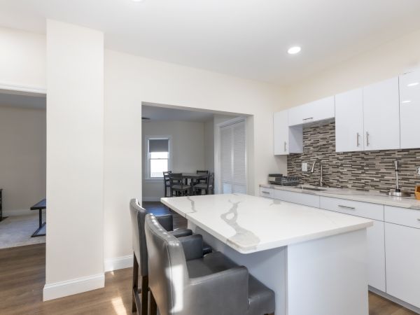 A modern kitchen with white cabinets, an island, gray chairs, and a tiled backsplash. A dining area is partially visible in the background.
