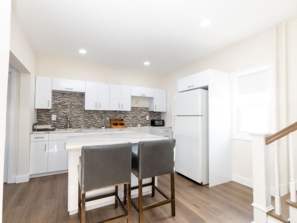A modern kitchen with white cabinets, backsplash, island, chairs, and a fridge, beside a staircase on wooden flooring.