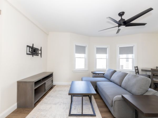 A minimalistic living room with a gray sofa, coffee table, TV mount, ceiling fan, and windows with light gray blinds.