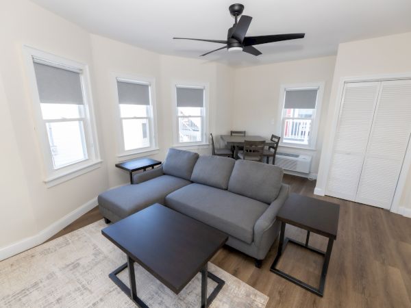 A modern, minimalist living room with a gray sectional sofa, dark wood tables, dining set, and a ceiling fan on wood flooring.
