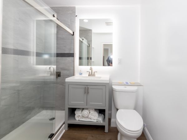 A modern bathroom with a glass shower, gray vanity, mirror, toilet, and rolled towels beneath the sink on wooden flooring.