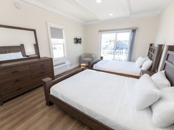 A bedroom with two wooden beds, a dresser with mirror, chair, window, sliding door to a balcony, and light-colored walls and flooring.