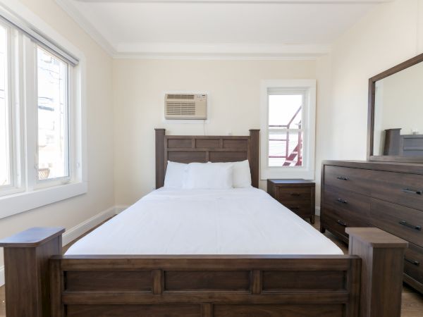 A simple bedroom with a wooden bed, white linens, wooden dresser, mirror, side tables, and an air conditioner on the wall.