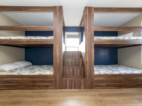 The image shows a wooden bunk bed setup with two levels on each side, separated by a central staircase, in a well-lit room.