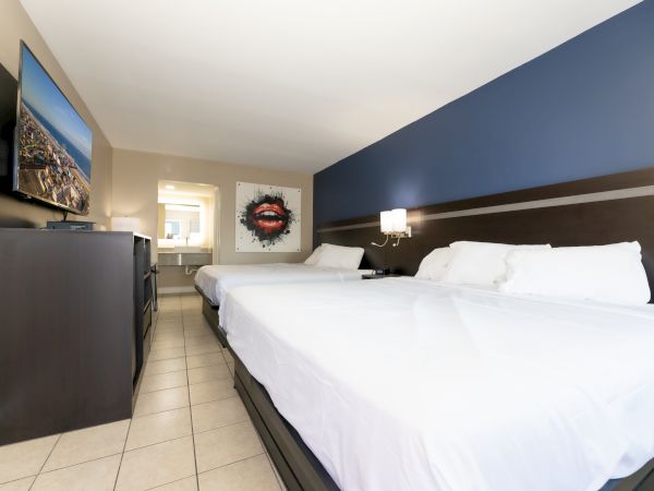 The image shows a hotel room with two beds, wall art of lips, a TV on a dresser, and a view into a bathroom area.
