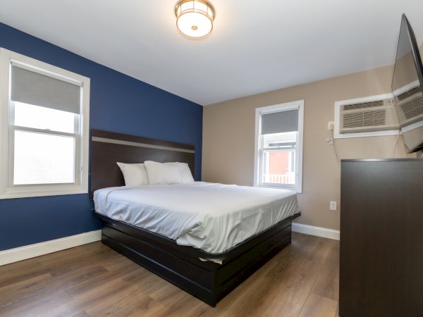 A modern bedroom features a neatly made bed, blue and beige walls, two windows, an air conditioning unit, and a wall-mounted TV.