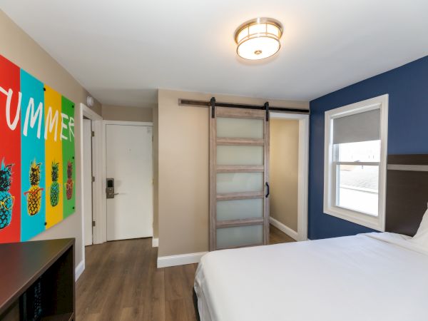 A modern room with a colorful "SUMMER" poster, sliding barn door, bed, and window.