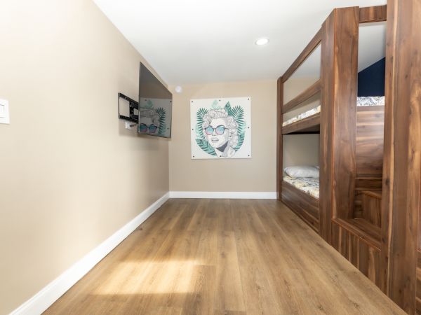 The image shows a room with wooden bunk beds, a mounted TV, and a wall art piece of a person wearing glasses with a leafy background.
